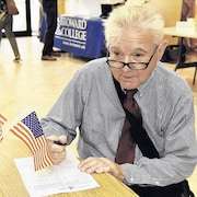 Rodney Nichols assis à un bureau remplit un document. Sur le bureau se trouvent des drapeaux américains.