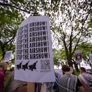 Une pancarte qui répète les mots « arrêtez le spectacle aérien » en avant-plan avec des manifestants derrière.