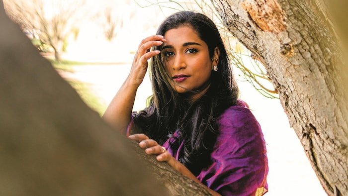 Une femme appuyée sur une branche d'arbre qui pose en regardant la caméra.