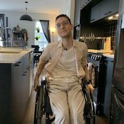 Un jeune homme est assis sur une chaise roulante. Il pose devant la caméra dans la cuisine d'une maison.