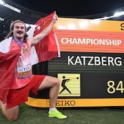Ethan Katzberg, portant sa médaille d'or et un drapeau canadien, lève le bras et sourit aux côtés du tableau indiquant son record de championnat à 84,70 m.