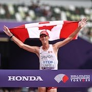 Evan Dunfee tient le drapeau du Canada au dessus de sa tête devant le dil d'arrivée de la course.